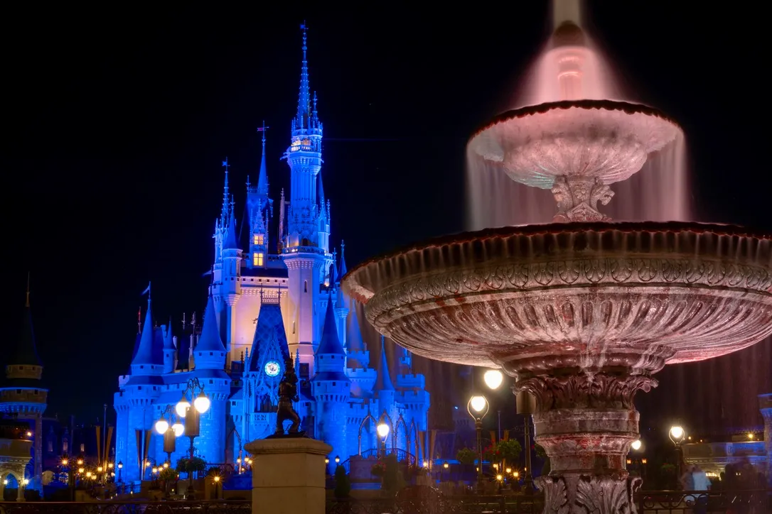 A fountain taken with long exposure in front of a floodlit blue castle at Walt Disney World Resort Magic Kingdom